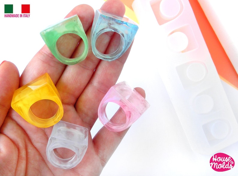 May include: Five colorful resin rings in a variety of shapes and sizes. The rings are made in a clear resin with different colors mixed in. The rings are displayed on a hand. The rings are made using a silicone mold that is also shown in the image. The mold has three different ring shapes. Handmade in Italy.