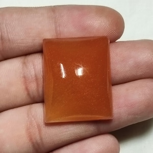 Könnte beinhalten: Ein rechteckiger, polierter Edelstein mit einer glatten Oberfläche im Cabochon-Stil. Der Stein weist einen leuchtenden, durchscheinenden Orangeton mit subtilem innerem Schimmer und Lichtreflexen auf seiner flachen, leicht gewölbten Oberseite auf.