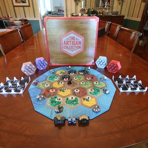 May include: A board game set featuring a hexagonal map with terrain tiles, small game pieces, and card holders. A large wooden box with a red frame displays the text THE ARTISAN COLLECTION in a hexagonal logo. Four small hexagonal containers are included.
