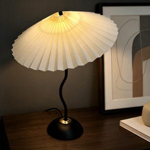 May include: A table lamp featuring a wide, off-white pleated fabric shade with a conical shape. The lamp has a slender, black, wavy metal stem that connects to a circular black base with a small gold-toned accent at the point of attachment.