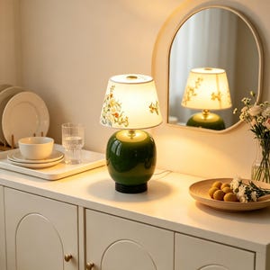 Könnte beinhalten: Eine Tischlampe mit einem glänzenden, dunkelgrünen Keramikfuß und einem weißen Stoffschirm mit zartem Blumenmuster. Die Lampe strahlt ein warmes Licht aus, das die Umgebung sanft erhellt, und steht auf einer ebenen Oberfläche.