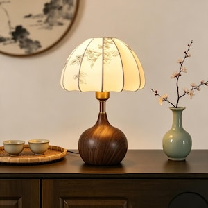 Könnte beinhalten: Eine Tischlampe mit einem dunkelbraunen Holzmaserungssockel und einem gewellten, cremeweißen Stoffschirm, der mit einer zarten grauen Blumenzeichnung versehen ist. Die Lampe hat einen messingfarbenen Metallhals, der den Sockel mit dem Schirm verbindet.