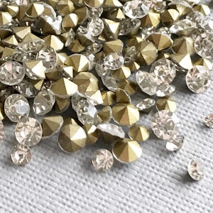 May include: A pile of small, sparkling rhinestones and gold-colored metal studs. The rhinestones are clear and faceted, while the studs have a pyramid shape. The items are scattered on a light gray surface, creating a shimmering effect.