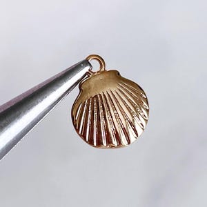 May include: A small, gold-colored seashell charm with detailed ridges, held by silver metal pliers against a soft, blurred background. The charm has a loop for attaching to jewelry. The overall theme is jewelry making and beach-themed accessories.