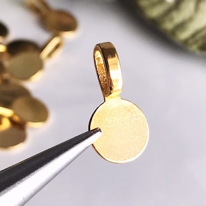 Puede incluir: Pendiente de metal dorado con forma de disco redondo y un bucle en la parte superior. El pendiente está siendo sujetado por unas pinzas.