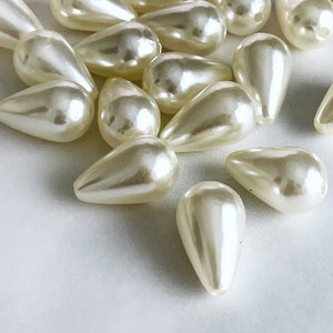 May include: A collection of off-white pearlescent teardrop-shaped beads. The beads are scattered on a white surface.