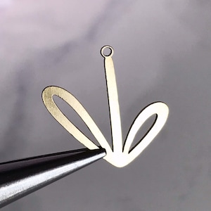 May include: A gold-colored metal charm in the shape of a stylized flower, held by tweezers. The charm has a central stem with a loop at the top and two curved petals on either side. The background is a soft, blurred mix of pink and gray.