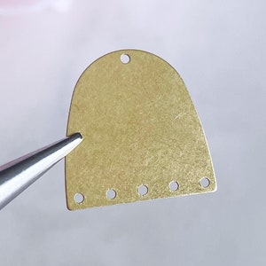 May include: A gold-colored metal pendant with a rounded top and a row of five holes along the bottom. The pendant has a small hole at the top for attaching a string or chain.