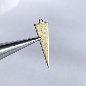 May include: A gold-toned triangle pendant with a loop at the top. The pendant is held by a pair of tweezers.