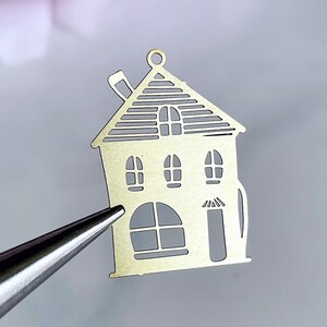 May include: Gold metal charm shaped like a house with a chimney, windows, and a door. The charm is cut out and has a textured surface.