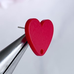 May include: A red heart-shaped earring with a small hole in the center. The earring is being held by a pair of pliers.