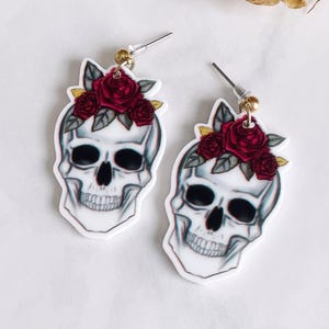 May include: A pair of white skull earrings with a red rose floral crown. The earrings have a silver post and a gold-colored bead. The skull has black eye sockets and a detailed jawline.