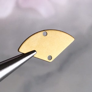 May include: Gold-colored metal half-circle charm with two holes. The charm is being held by a pair of tweezers.