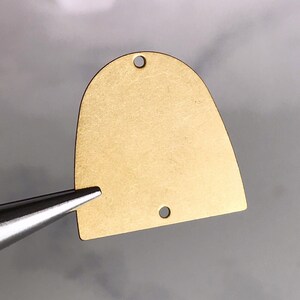 May include: A gold-colored metal pendant shaped like a rounded rectangle with two holes at the top. The pendant is smooth and has a slightly textured surface.