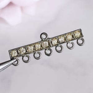 May include: A silver-toned jewelry component with a rectangular bar adorned with small, clear rhinestones. The bar has a series of small loops along the bottom edge and a single loop at the top. The component is held by a pair of tweezers.