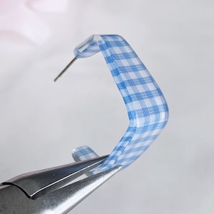 May include: A pair of blue and white gingham patterned hoop earrings. The earrings are being held by a pair of pliers.