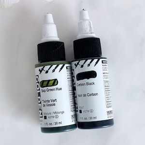 Puede incluir: Dos botellas de pintura acrílica Golden, una etiquetada como "Sap Green Hue" y la otra como "Carbon Black". Cada botella contiene 30 ml. Las botellas tienen tapas negras y etiquetas blancas con texto negro.