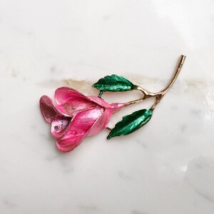 May include: A pink enamel rose brooch with green enamel leaves. The rose is stylized and has a gold tone finish.