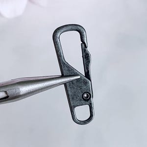 May include: A close-up of a gunmetal-colored metal clasp. The clasp has a rectangular body with a rounded loop at the bottom and a spring-loaded hook at the top. A pair of metal pliers holds the clasp.