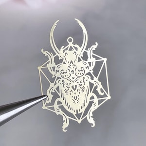 Puede incluir: Un colgante de metal dorado detallado en forma de escarabajo estilizado, sujeto con pinzas. El escarabajo está intrincadamente diseñado con patrones ornamentados y está encerrado dentro de un marco geométrico. El colgante mide aproximadamente 5 cm de largo.