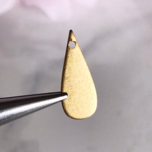 May include: A small, teardrop-shaped brass pendant with a matte finish. The pendant has a small hole at the top for stringing. Held by a pair of silver tweezers against a soft, blurred background.