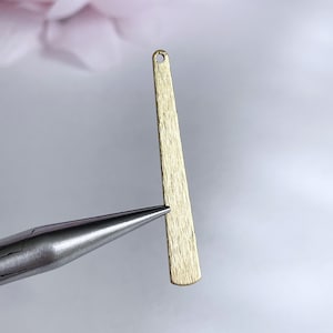 May include: A gold-colored metal pendant with a textured surface and a rounded top. The pendant has a small hole at the top for attaching a chain or string.