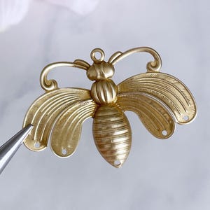 Op de afbeelding: Een goudkleurige metalen bijenbedel met gedetailleerde vleugels en lichaam. De bij heeft een lus aan de bovenkant om op te hangen en kleine gaatjes om te bevestigen. De vleugels hebben een gebogen ontwerp en het lichaam is geribbeld.