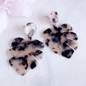 Puede incluir: Un par de pendientes colgantes con forma de hoja y estampado carey en tonos crema, marrón y negro. Los pendientes están hechos de un material ligero y tienen cierre de poste para orejas perforadas.