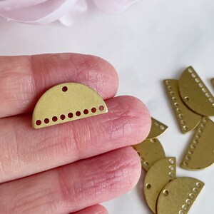 May include: A close-up of a gold-toned metal half-circle charm with a hole at the top and a row of small holes along the bottom edge. The charm is being held in a hand, and there are several other similar charms in the background.