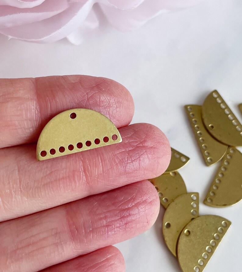 May include: A close-up of a gold-toned metal half-circle charm with a hole at the top and a row of small holes along the bottom edge. The charm is being held in a hand, and there are several other similar charms in the background.