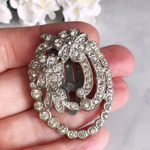 May include: A silver hair clip with a floral design and clear rhinestones. The clip is oval shaped and has a row of rhinestones around the edge.