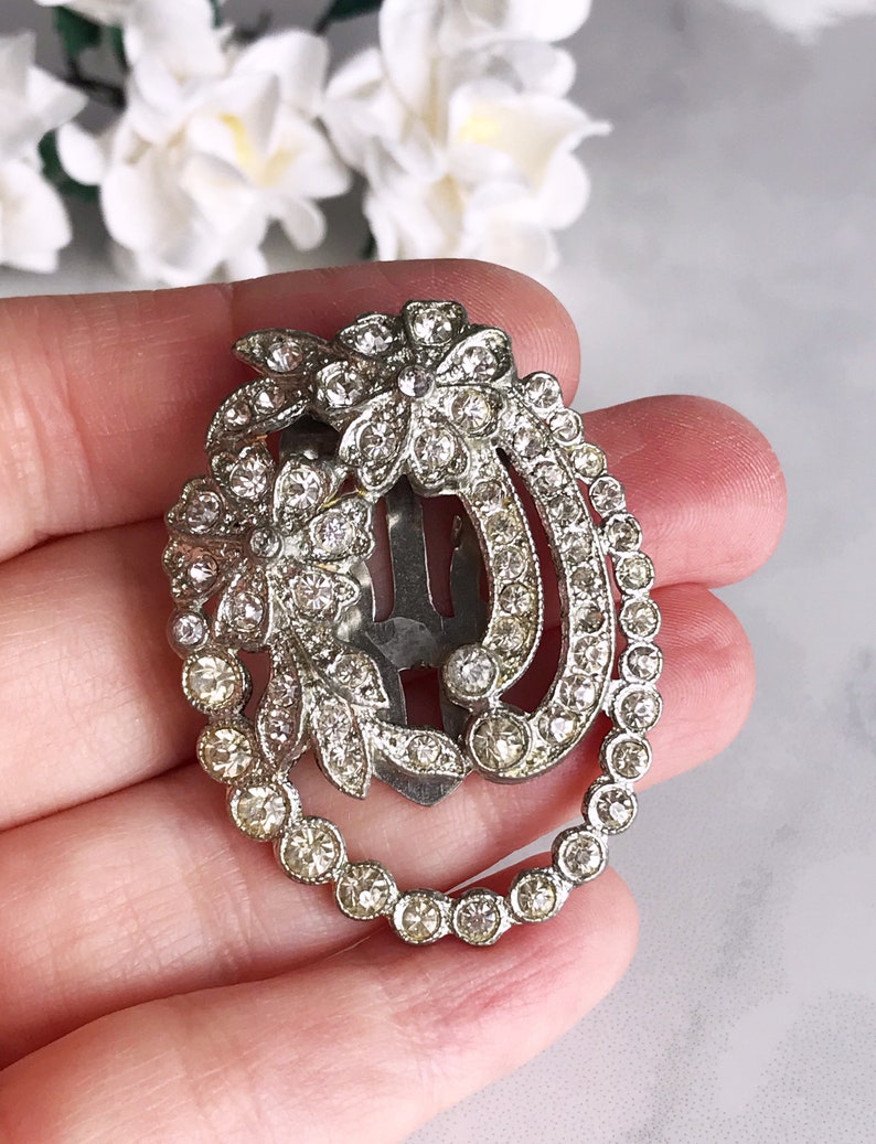 May include: A silver hair clip with a floral design and clear rhinestones. The clip is oval shaped and has a row of rhinestones around the edge.