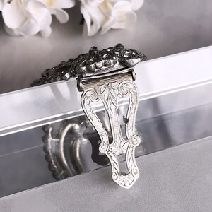 May include: Silver-toned metal clasp with an ornate design. The clasp is decorated with a floral pattern and has a hinged closure.