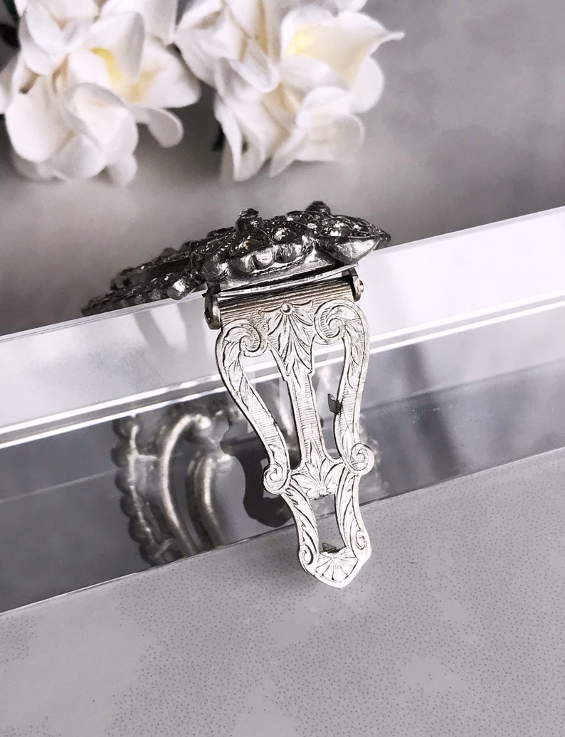May include: Silver-toned metal clasp with an ornate design. The clasp is decorated with a floral pattern and has a hinged closure.