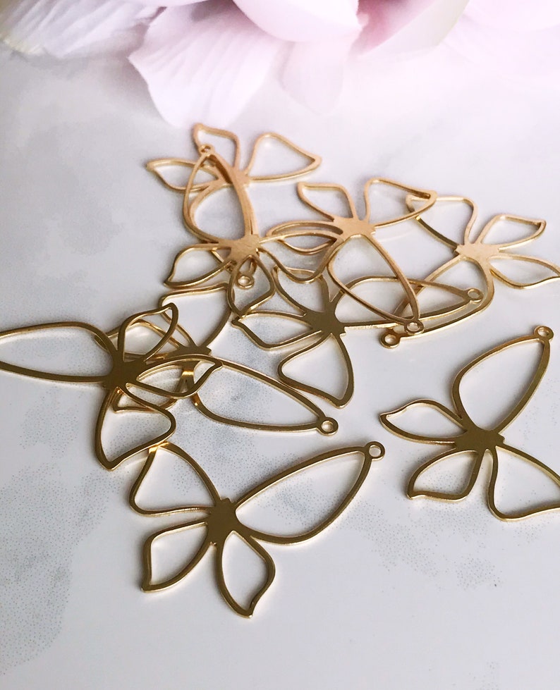 May include: A set of six gold-colored metal butterfly charms with openwork designs. The charms have a loop at the top for attaching to jewelry or other crafts.