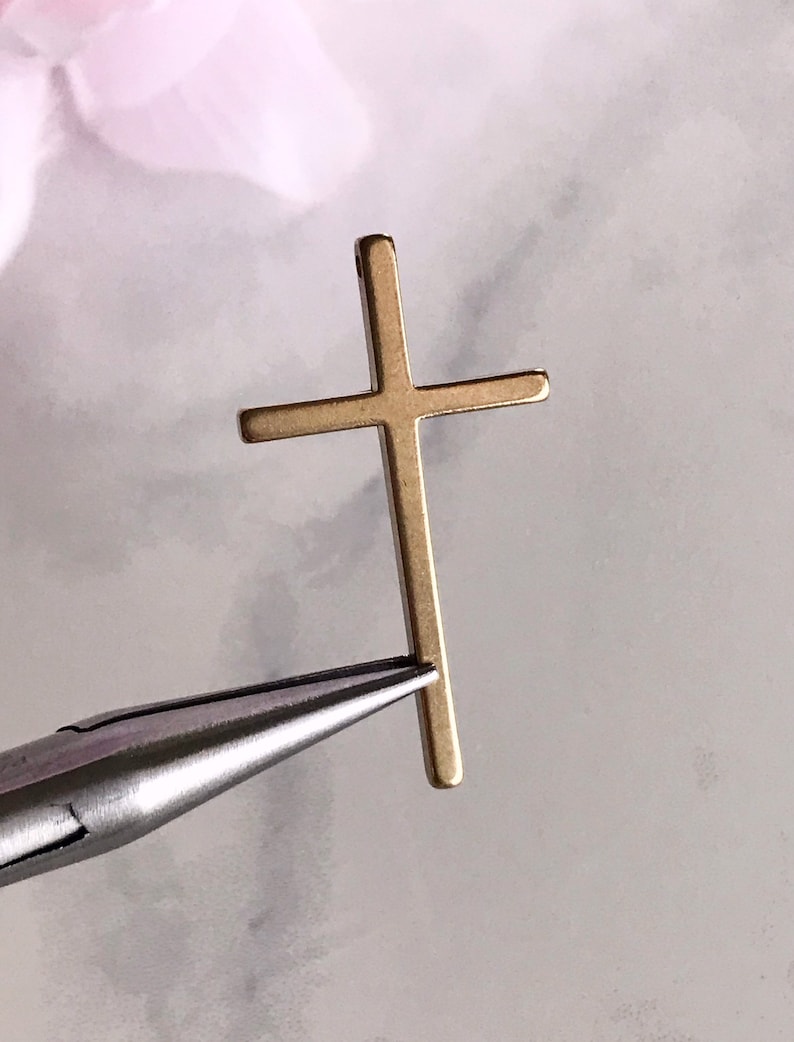 May include: A gold-colored metal cross pendant with a simple design. The cross is being held by a pair of pliers.
