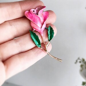 May include: A pink enamel rose with green enamel leaves and a gold stem. The rose is a vintage charm or pendant.