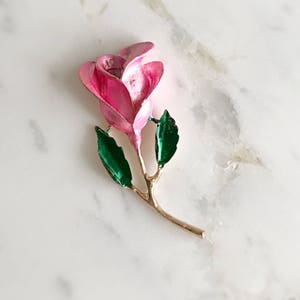 May include: A pink enamel rose brooch with green enamel leaves and a gold stem.