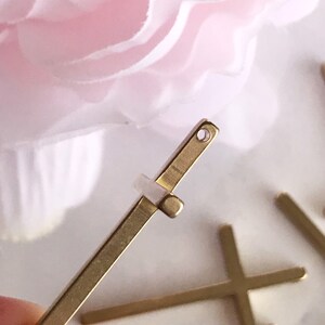 May include: Gold metal cross charm with a small hole at the top. The charm is flat and has a simple design.