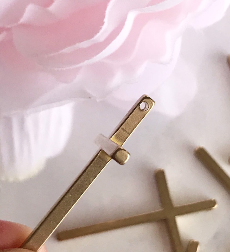 May include: Gold metal cross charm with a small hole at the top. The charm is flat and has a simple design.