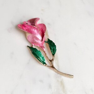 May include: A pink enamel rose brooch with green enamel leaves and a gold tone stem.