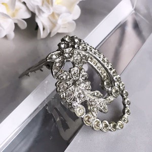 May include: A silver-toned brooch with a floral design, featuring a large oval shape with a smaller oval cutout in the center. The brooch is adorned with clear rhinestones.