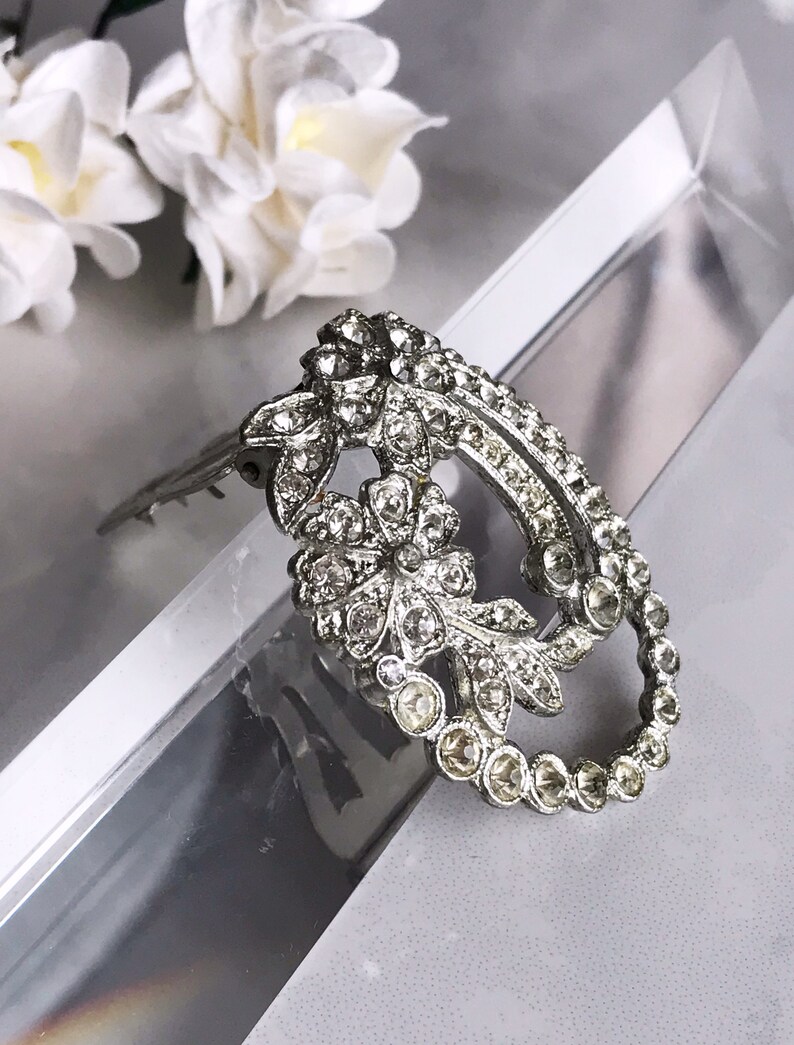 May include: A silver-toned brooch with a floral design, featuring a large oval shape with a smaller oval cutout in the center. The brooch is adorned with clear rhinestones.