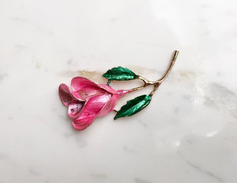 May include: A pink enamel rose brooch with green enamel leaves. The rose is stylized and has a gold tone finish.