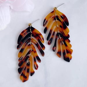 May include: A pair of leaf-shaped earrings in a tortoiseshell pattern. The earrings are made of a translucent material with black, brown, and yellow hues. They are linked by small gold-coloured chains and have silver-coloured posts.
