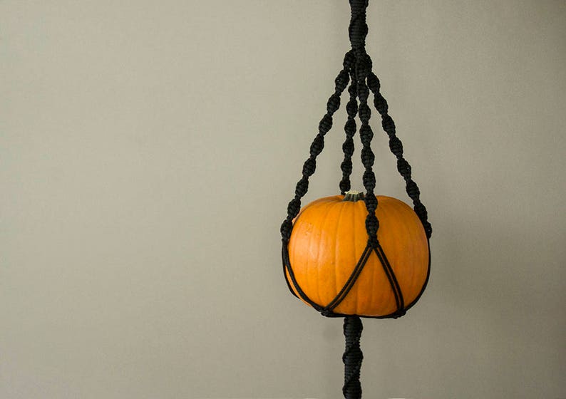 May include: A black macrame plant hanger with a small orange pumpkin hanging inside.