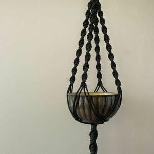 May include: Black macrame plant hanger with a brown ceramic pot. The hanger has a long tassel at the bottom.
