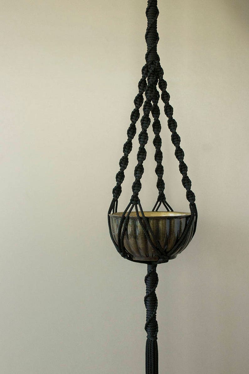 May include: Black macrame plant hanger with a brown ceramic pot. The hanger has a long tassel at the bottom.