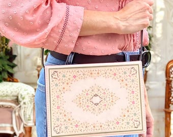 May include: A rectangular crossbody bag featuring a light pink surface with a detailed, symmetrical floral pattern in shades of pink, green, and brown. The bag has a dark brown strap and is worn over a pink long-sleeved shirt and blue denim jeans.