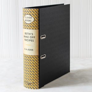 May include: A black ring binder with silver metal clasps. The spine displays a tan label with the text "CLEVER BOOKS" and "BETH'S BAKE OFF RECIPES" in black. The spine has a yellow and black geometric pattern.